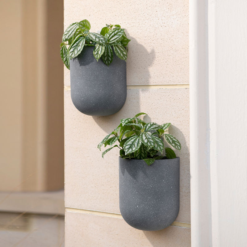 Two weathered gray planters are hung on a beige outer wall, both potted with green plants.
