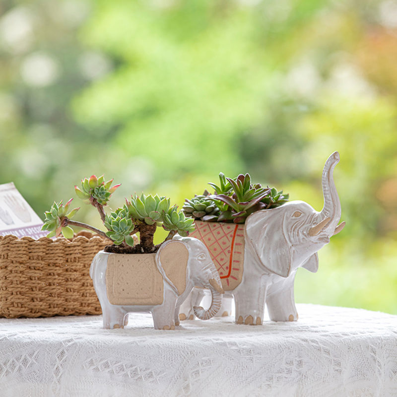 The set of two elephant planters with succulents in them is displayed on an outdoor table, in front of a basket.  