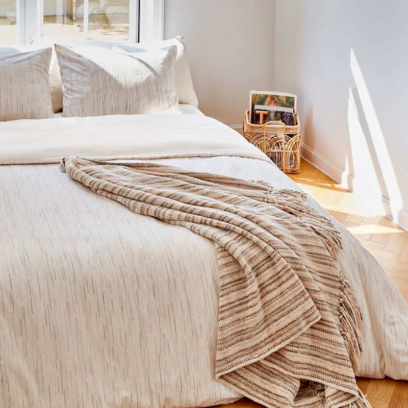 The woven chenille throw blanket is placed on a light-colored bed, showing its subtle brown stripes.