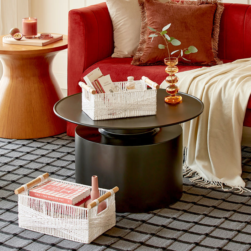 A large white basket is placed on a plaid carpet while a smaller one is set on a black table, both in front of a red sofa.