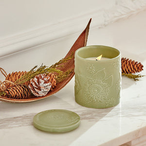 A burning jar of wooden pine scented candle is placed on a marble table, surrounded by pine cones.