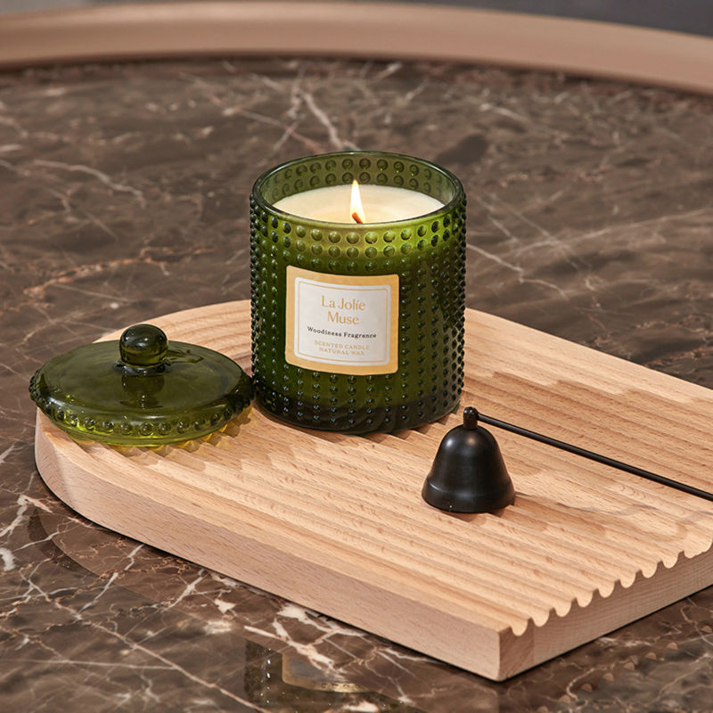 A burning candle in a transparent green jar, placed on a wooden tray with a glass lid and a candle snuffer next to it.