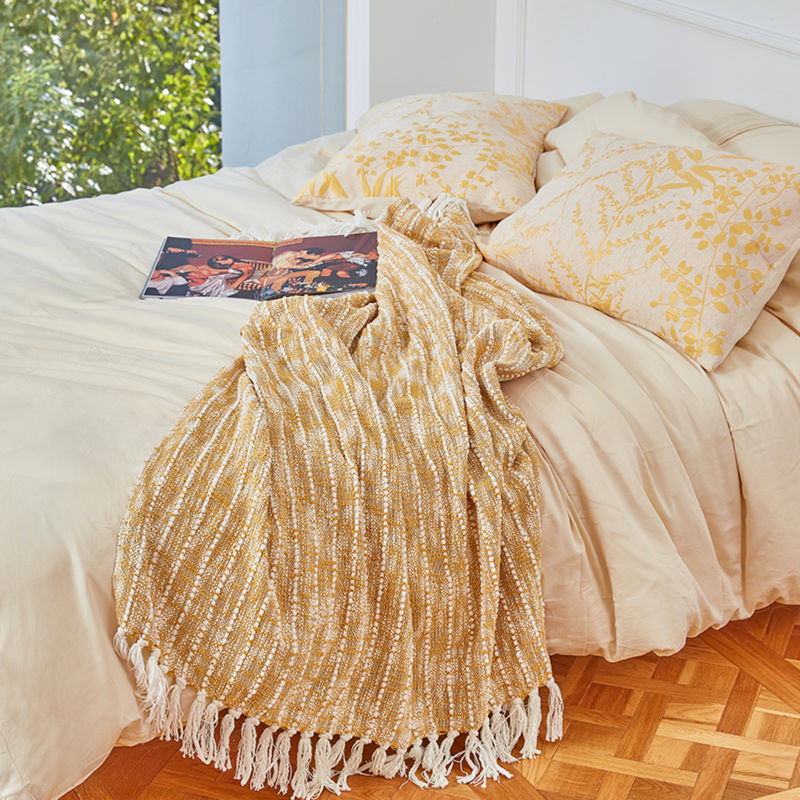 A yellow and white cotton throw is placed on a beige color bed while a poster is placed on top of the blanket.